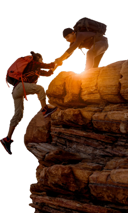 Two outdoor climbers navigating rocky terrain: one climber positioned higher on a rock formation reaches down to assist another climber in ascent. Both wearing backpacks and adventure-ready clothing, the scene is illuminated by warm light, suggesting sunrise or sunset. This image embodies teamwork, perseverance, and the spirit of adventure in nature.