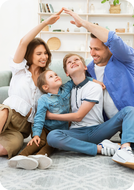 Cheerful family moment indoors: a woman in a white blouse and a man in a blue jacket joyfully encircle their two children, a boy with light brown hair in a light-colored shirt and a girl in a denim outfit. The adults playfully form an arch with their hands, creating a nurturing atmosphere. Seated on a soft rug in a cozy living room filled with plants and shelves, this image radiates joy, love, and strong familial bonds.